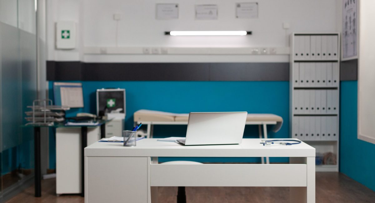 Medical consultation room with white desk, laptop, stethoscope, and examination table against teal walls